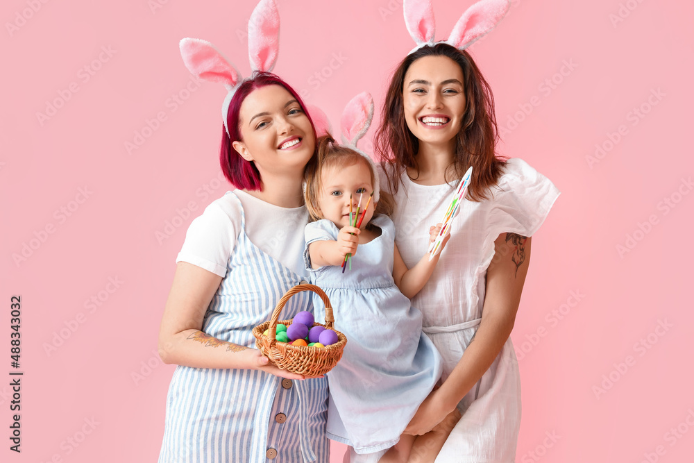 Young lesbian couple with little daughter, Easter eggs, brushes and paints on color background