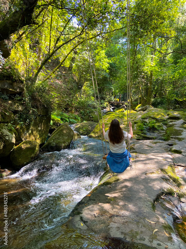 Young beautiful woman swinging on a wooden swing hanging by rope on a tree branch over Sampaio river in Cinfaes, near the village of Mourilhe, Portugal. Girl having fun on a swing underneath a tree.