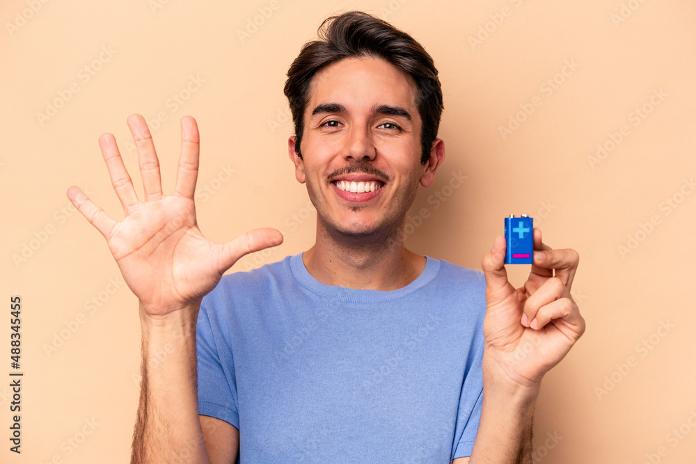 Young caucasian man holding a batterie isolated on beige background smiling cheerful showing number five with fingers. Young caucasian man holding a batterie isolated on beige background smiling cheerful showing number five with fingers.
