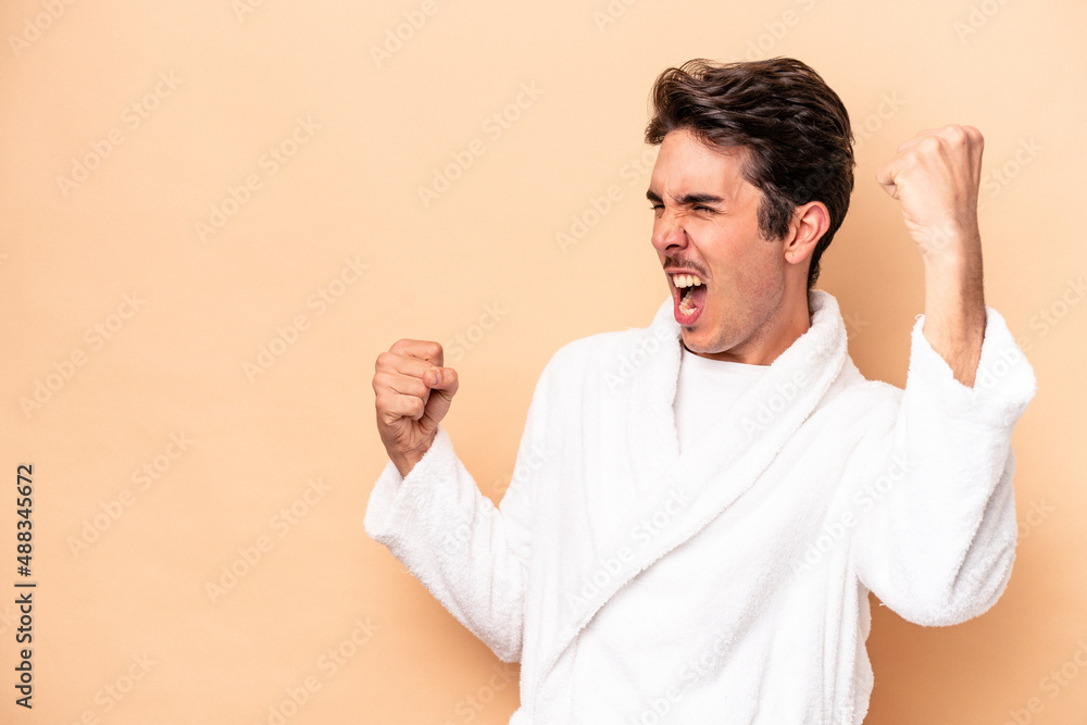 Young caucasian man wearing a bathrobe isolated on beige background raising fist after a victory, winner concept. Young caucasian man wearing a bathrobe isolated on beige background raising fist after a victory, winner concept.