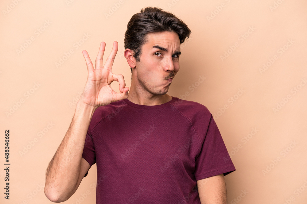 Young caucasian man isolated on beige background winks an eye and holds an okay gesture with hand. Young caucasian man isolated on beige background winks an eye and holds an okay gesture with hand.