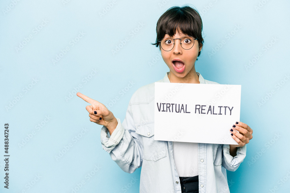 Young hispanic woman holding a virtual reality placard isolated on blue background pointing to the side Young hispanic woman holding a virtual reality placard isolated on blue background pointing to the side