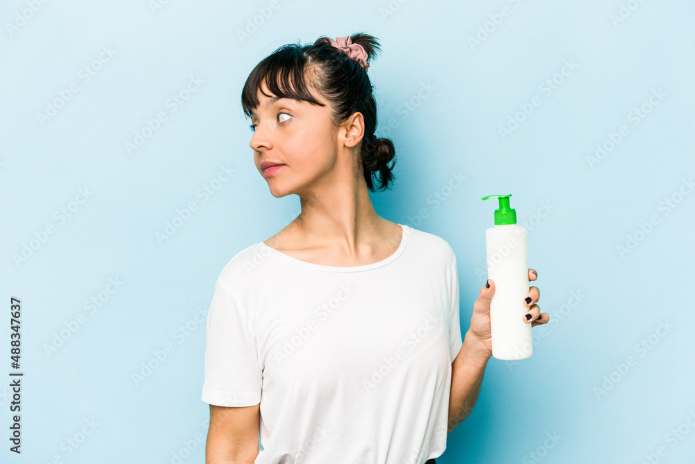 Young hispanic woman holding a body lotion isolated on blue background looks aside smiling, cheerful and pleasant. Young hispanic woman holding a body lotion isolated on blue background looks aside smiling, cheerful and pleasant.