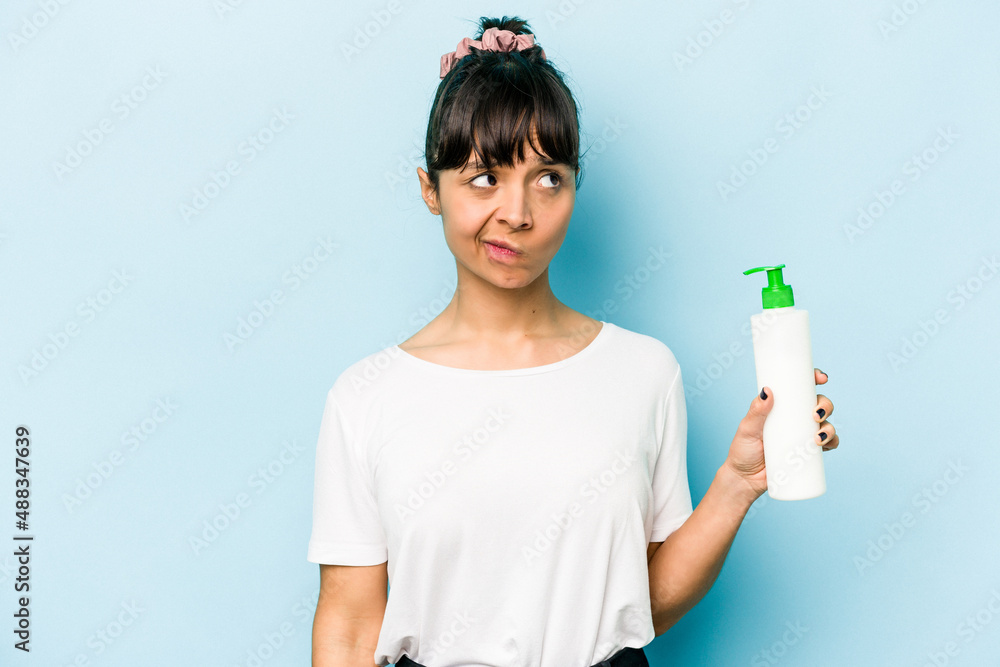 Young hispanic woman holding a body lotion isolated on blue background confused, feels doubtful and unsure. Young hispanic woman holding a body lotion isolated on blue background confused, feels doubtful and unsure.