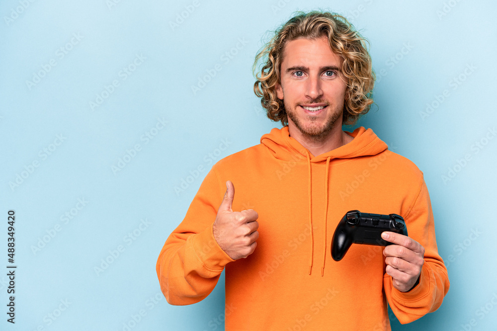 Young caucasian man playing with a video game controller isolated on blue background smiling and raising thumb up Young caucasian man playing with a video game controller isolated on blue background smiling and raising thumb up
