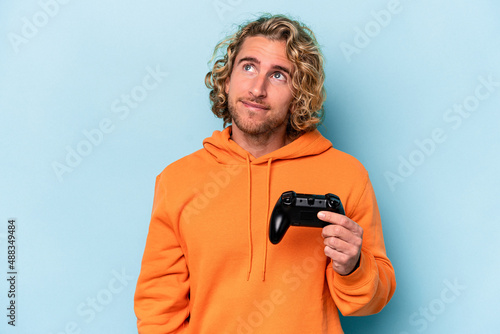 Young caucasian man playing with a video game controller isolated on blue background dreaming of achieving goals and purposes