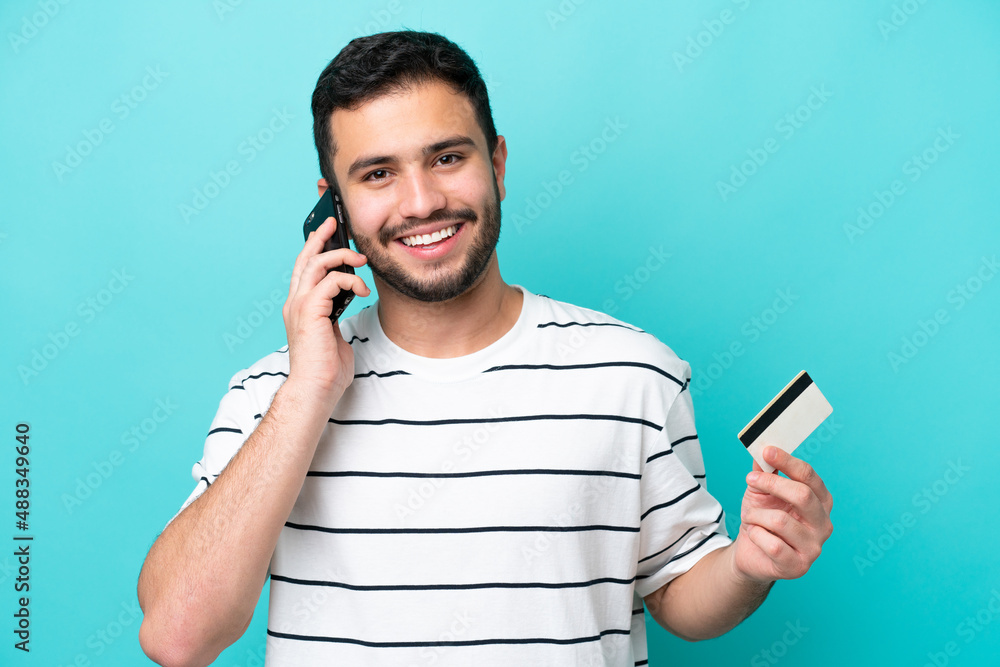 © luismolinero - Young Brazilian man isolated on blue background keeping a conversation with the mobile phone and holding a credit card