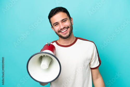 Young Brazilian man isolated on blue background holding a megaphone and smiling a lot