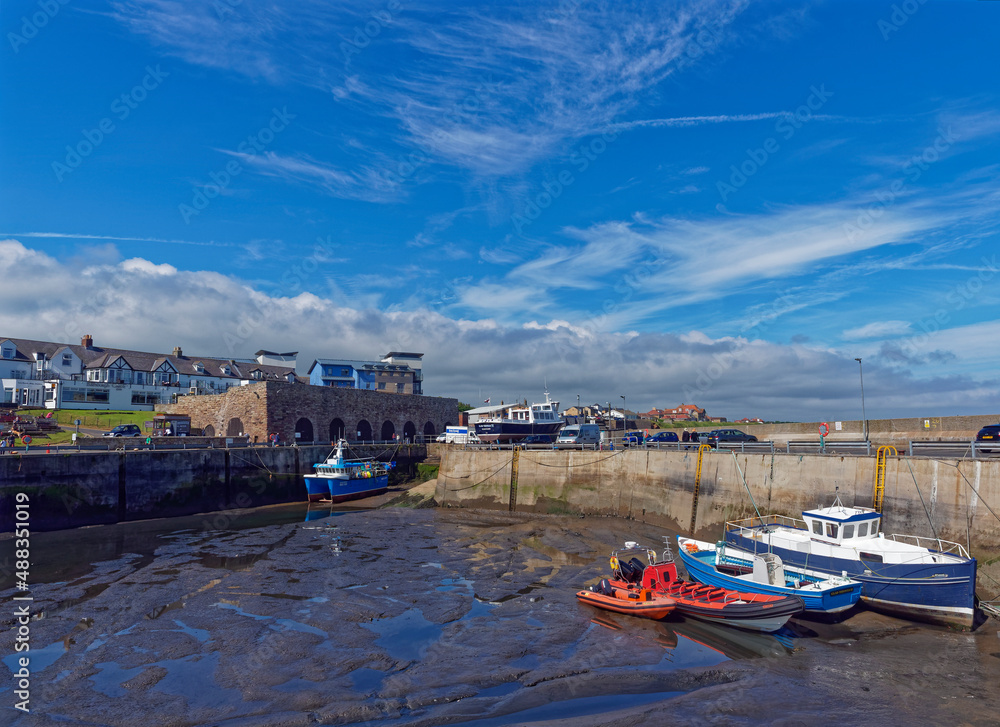 North Sunderland harbour at Seahouses at Low Tide, with Boats resting on the silty bottom of the Harbour, overlooked by Houses and Commercial Buildings.