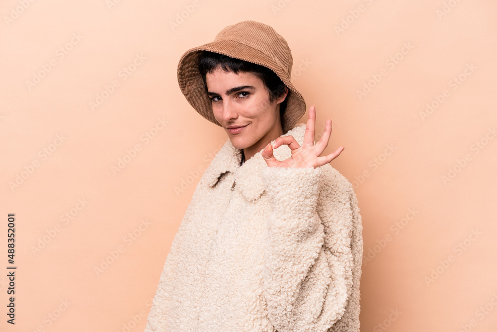 Young caucasian woman isolated on beige background winks an eye and holds an okay gesture with hand.