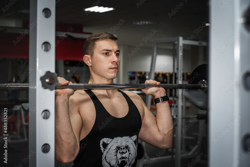 Fototapeta premium Athletic muscled young man in black t-shirt lifting barbell among sport gym stuff on black grey dark background