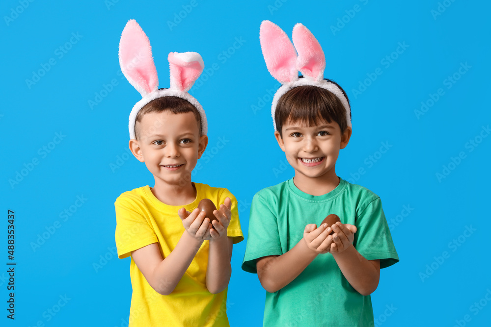 Cute little boys with bunny ears and chocolate Easter eggs on blue background Cute little boys with bunny ears and chocolate Easter eggs on blue background