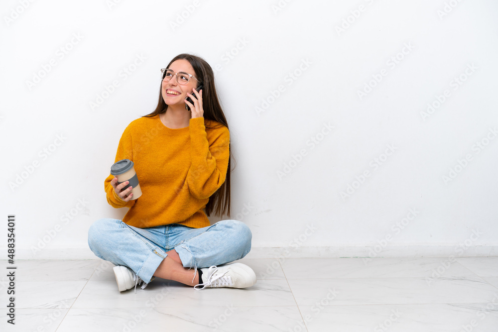 Young girl sitting on the floor isolated on white background holding coffee to take away and a mobile