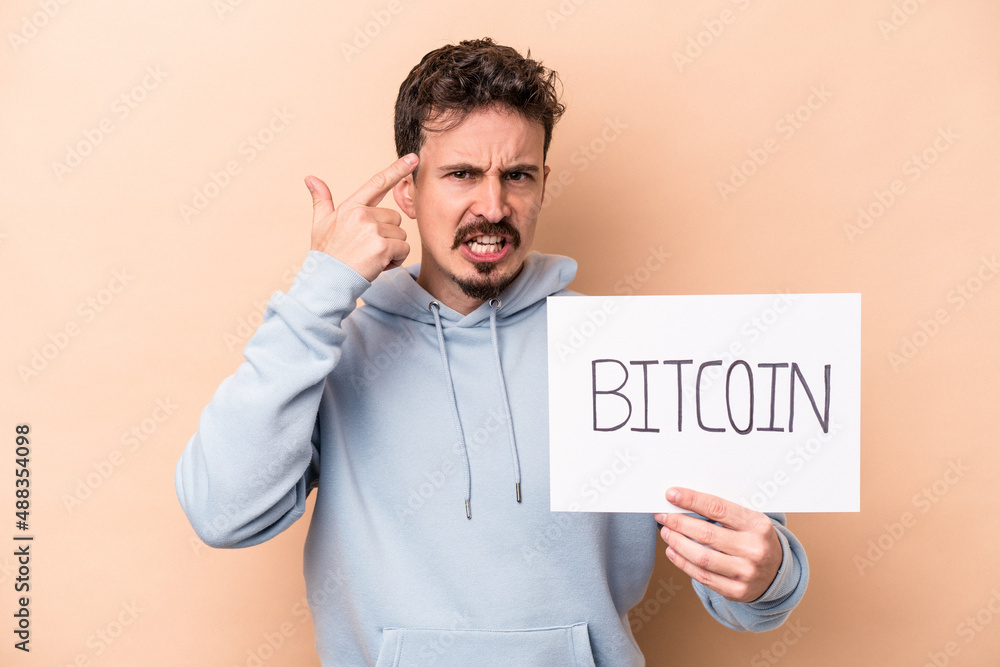 Young caucasian man holding a bitcoin placard isolated on beige background showing a disappointment gesture with forefinger. Young caucasian man holding a bitcoin placard isolated on beige background showing a disappointment gesture with forefinger.