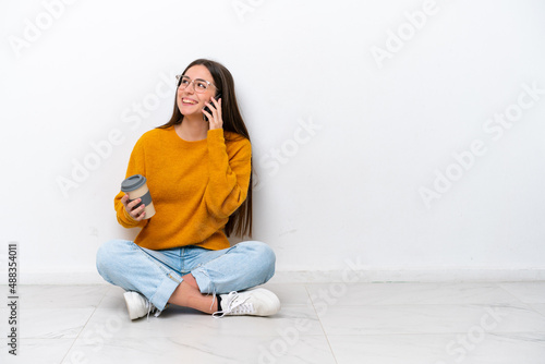 Young girl sitting on the floor isolated on white background holding coffee to take away and a mobile