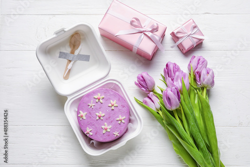 Plastic lunch box with tasty bento cake and flowers on light wooden background
