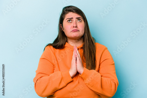 Canvastavla Young caucasian overweight woman isolated on blue background holding hands in pray near mouth, feels confident