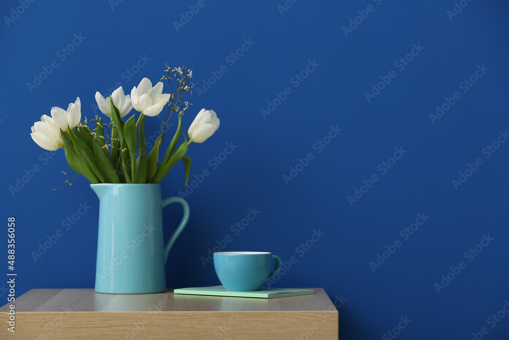 Jug with tulips, cup and notebook on table near color wall