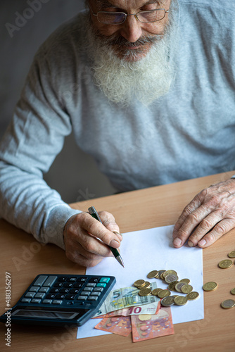 Old bearded senior man with calculator and bills counting euro money and writing notes on white sheet of paper.