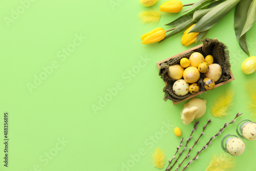 Box with painted Easter eggs, tulip flowers and willow branches on green background
