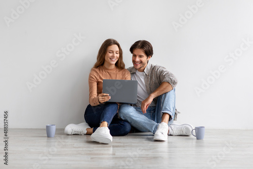 Glad smiling young european man and lady look at laptop planning new interior, sitting on floor in empty room