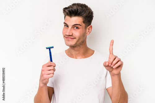 Young caucasian man shaving his beard isolated on white background showing number one with finger.