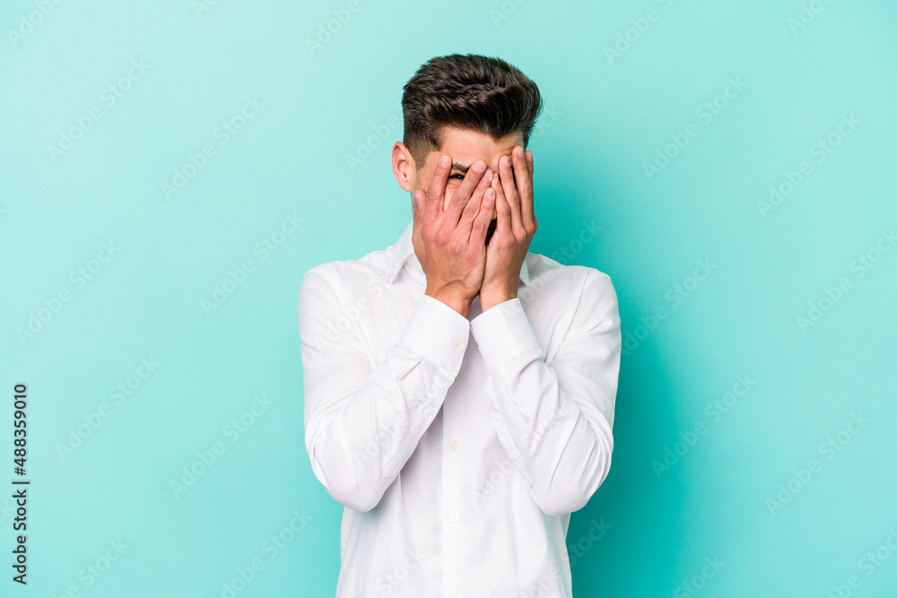 Young caucasian man isolated on blue background blink at the camera through fingers, embarrassed covering face. Young caucasian man isolated on blue background blink at the camera through fingers, embarrassed covering face.