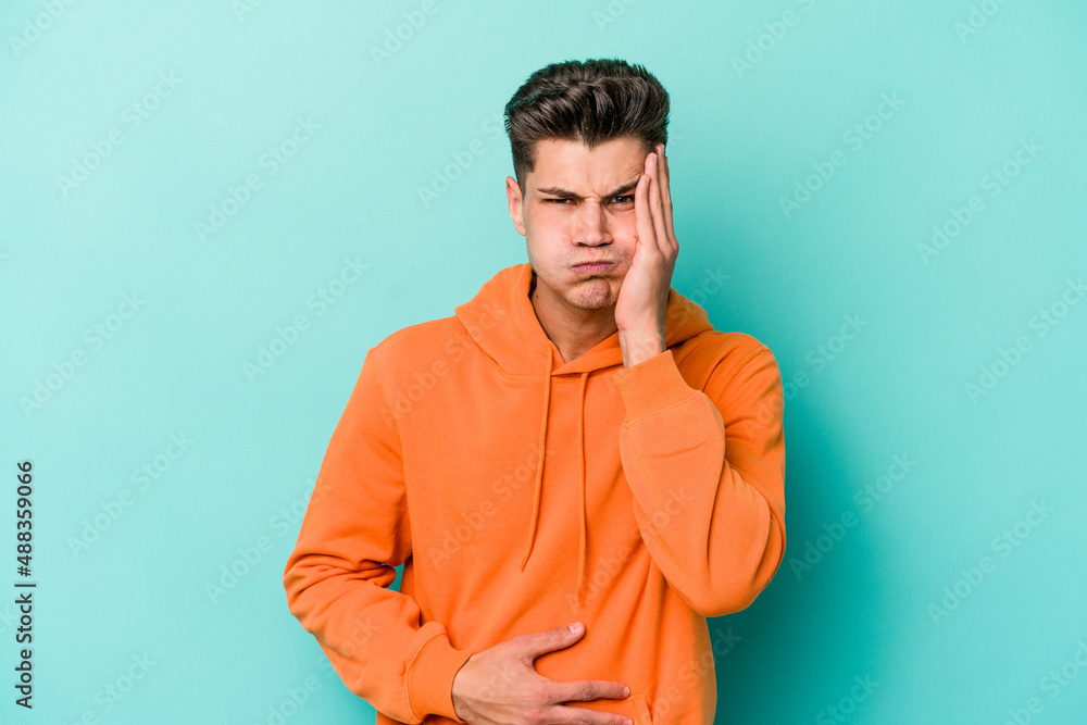 Young caucasian man isolated on blue background blows cheeks, has tired expression. Facial expression concept.