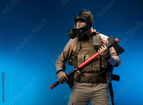 a soldier in military clothes with a bulletproof vest in a gas mask holds a red fire axe in his hands