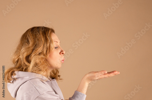 Woman with red curly hair blows on the palm of her hand sends an air kiss. Studio portrait on beige monochrome background