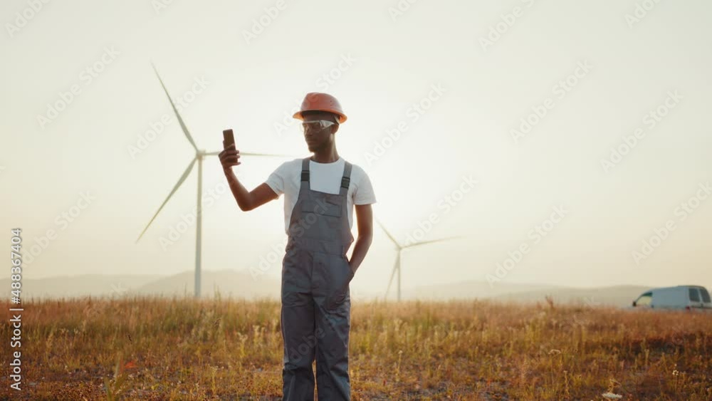 Engineer in uniform using modern smartphone for video call among windmill farm. Concept of people, technology and green energy. Engineer having video call on mobile among windmill farm. Engineer in uniform using modern smartphone for video call among windmill farm. Concept of people, technology and green energy. Engineer having video call on mobile among windmill farm.