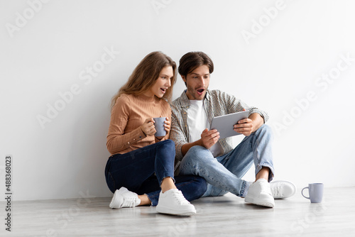 Surprised millennial caucasian male and female with open mouths look at tablet sit on floor in empty room