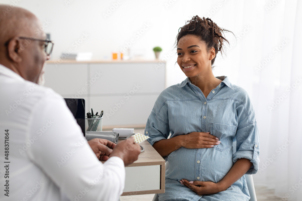 Fototapeta premium Mature experienced black doctor showing pills to pregnant lady