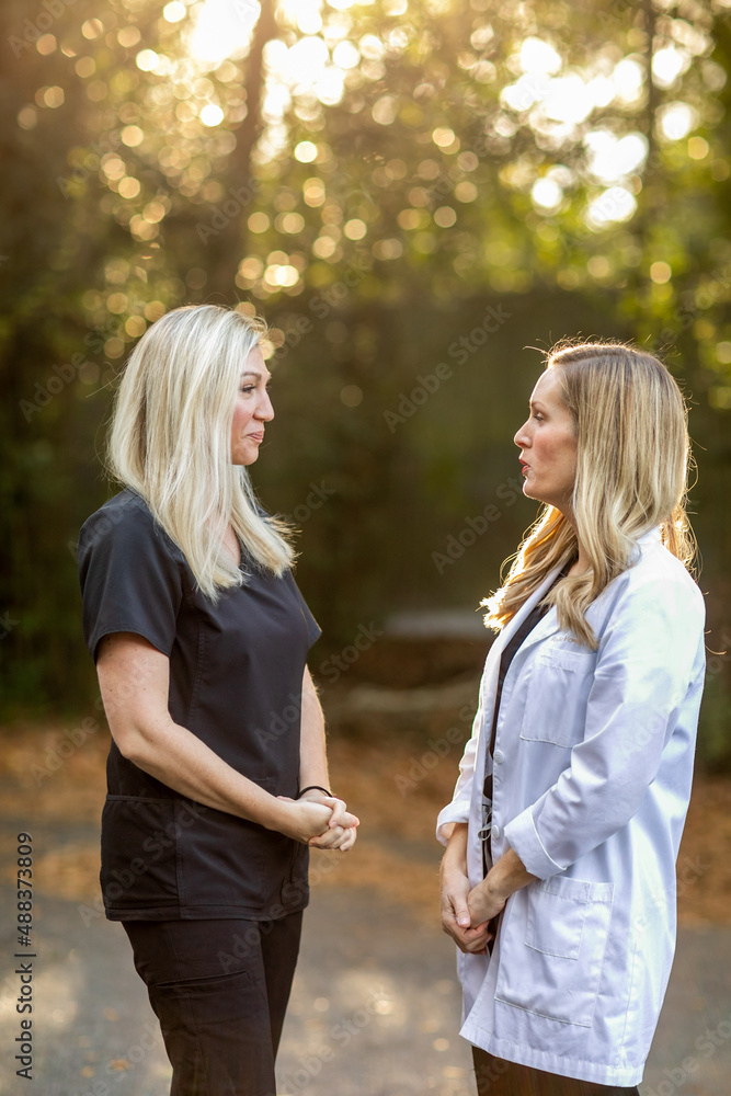 Fototapeta premium Two female medical professionals in black scrubs and one is wearing a white lab coat standing outside talking