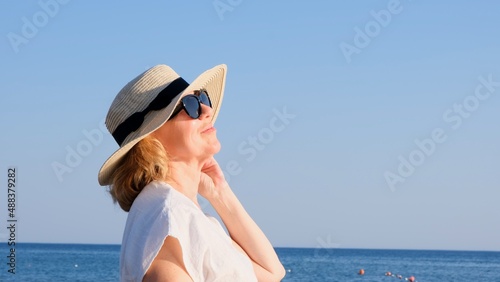 Portrait of a contented 50 year old woman wearing a straw hat and sunglasses enjoying the sun against the blue sea. Summer, vacation, vacation, active retirees