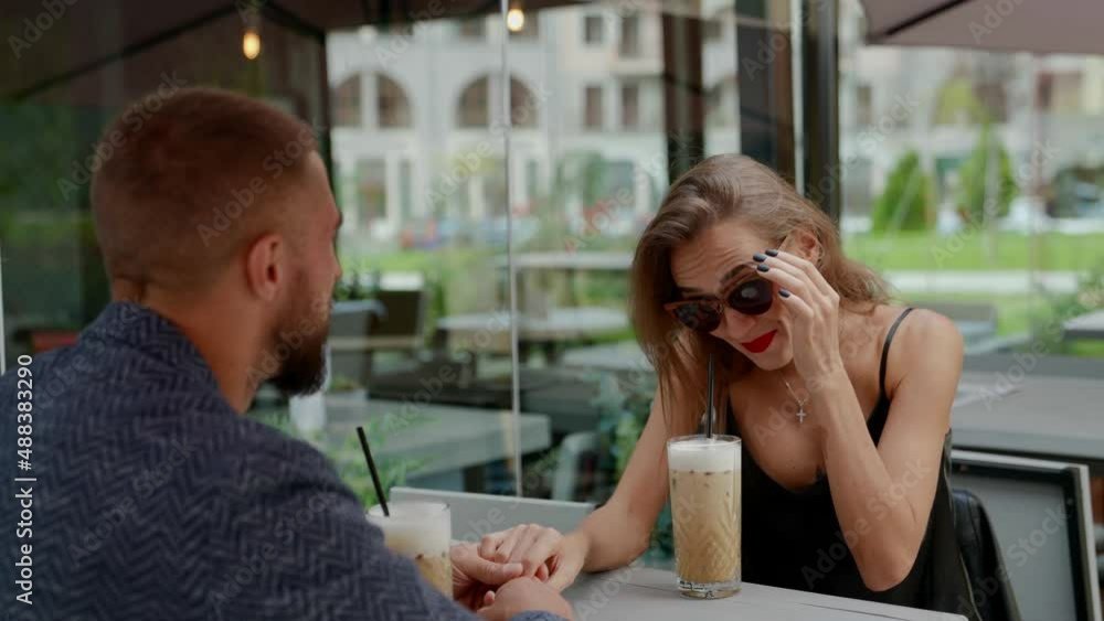 a couple is sitting at a table in an outdoor cafe. a blonde with bright lipstick drinks a cocktail and takes off sunglasses and smiles. the man is visible from the back. the camera is moving a couple is sitting at a table in an outdoor cafe. a blonde with bright lipstick drinks a cocktail and takes off sunglasses and smiles. the man is visible from the back. the camera is moving