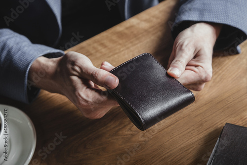 A business man at a wooden table holds a leather wallet in a cafe and waits for a lunch bill. No face, close-up