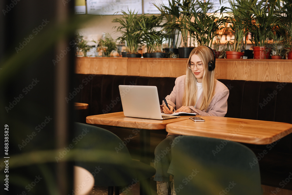 A young millennial freelancer woman with headphones works and studies online using a mobile phone and wireless technologies while sitting in the workplace. A business woman takes notes in a planner A young millennial freelancer woman with headphones works and studies online using a mobile phone and wireless technologies while sitting in the workplace. A business woman takes notes in a planner