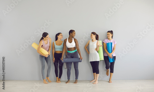 Group of smiling girls in modern sports outfits and with workout mats standing in fitness studio. Five happy fit sporty beautiful young multiethnic women in leggings and yoga pants posing by gym wall