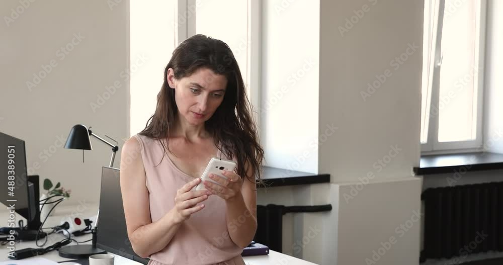 Young 35s businesswoman standing alone at workplace with smartphone, read message, learn received information, solve business distantly using modern tech, internet connection. Mobile app usage concept Young 35s businesswoman standing alone at workplace with smartphone, read message, learn received information, solve business distantly using modern tech, internet connection. Mobile app usage concept