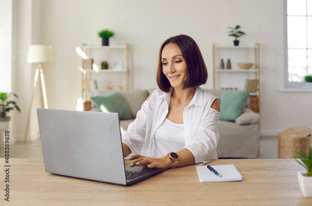 Beautiful smiling business lady working remotely in her home office using laptop. Middle-aged woman sitting at table in front of laptop and notebook and looking for useful information on Internet. Beautiful smiling business lady working remotely in her home office using laptop. Middle-aged woman sitting at table in front of laptop and notebook and looking for useful information on Internet.
