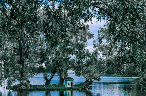 A small island with silvery willows in the midst of a beautiful lake against the blue sky and the Gatchina Palace near Saint Petersburg.	