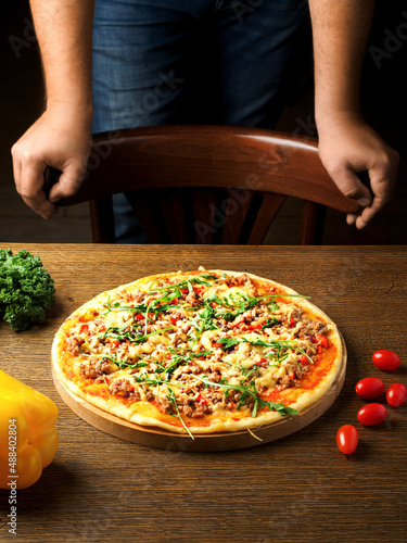 Pizza with minced meat, cheese, vegetables and arugula on a wooden table. Male hands in the frame. Copy space. Close up. Man is going to eat pizza.