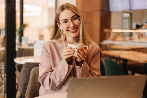 Portrait of a smiling student in headphones drinking coffee and working using a mobile phone and laptop in a cafe. A young business woman freelancer with glasses works remotely online from coffee shop