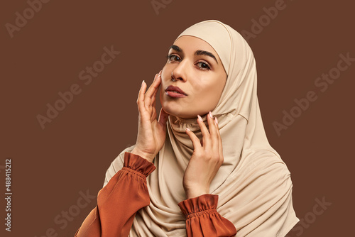 Portrait of beautiful modern muslim woman with natural make-up dressed in beige hijab posing on brown background in studio. Facial skin care, female beauty. photo