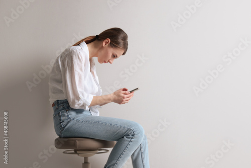 Woman with bad posture using smartphone while sitting on stool against light grey background, space for text