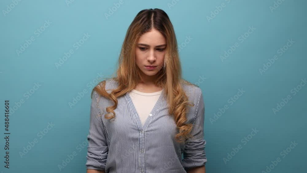 Portrait of upset woman with wavy hair expressing negative emotions and sorrow, has bad mood, being sad and unhappy, wearing striped shirt. Indoor studio shot isolated on blue background. Portrait of upset woman with wavy hair expressing negative emotions and sorrow, has bad mood, being sad and unhappy, wearing striped shirt. Indoor studio shot isolated on blue background.