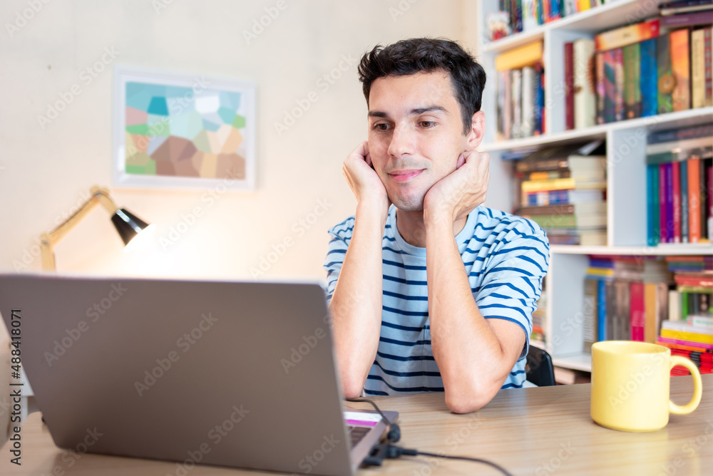 Student sitting at desk in his bedroom and using laptop to study, with book and paper by his side. Distance learning or e-learning through digital platforms. Student sitting at desk in his bedroom and using laptop to study, with book and paper by his side. Distance learning or e-learning through digital platforms.