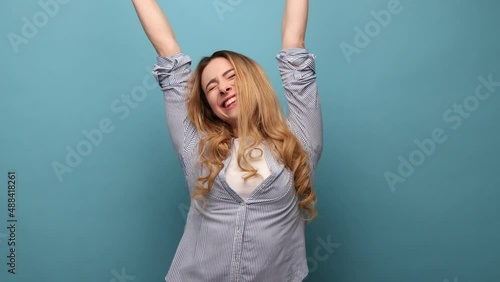 Portrait of victorious delighted happy woman raising hands, shouting for joy, screams celebrating win success, thrilled emotions, wearing striped shirt. Indoor studio shot isolated on blue background.