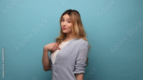 This is me. Portrait of serious young woman with wavy hair standing, pointing herself with proud face and looking at camera, wearing striped shirt. Indoor studio shot isolated on blue background. photo